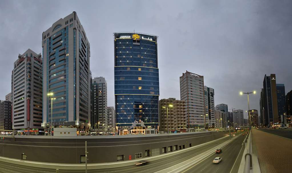 Hotel Jannah Burj Al Sarab Abu Dhabi Esterno foto The photo depicts a modern urban landscape featuring several high-rise buildings. In the foreground, there is a road with vehicles, suggesting a busy city environment. Prominent among the buildings is a tall, blue structure with a distinctive logo at